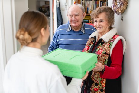 Frau überreicht eine grüne Steroporbox an älteres Paar an der Haustür