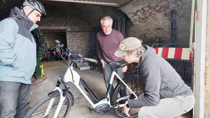 Bild mit drei Personen, zwei Männer arbeiten an einem Fahrrad, ein Mann mit Fahrradhelm steht daneben