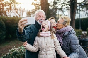 Großvater nimmt ein Selfie einer Gruppe mit Mobiltelefon in einem Park auf an einem Wintertag auf. Die Großmutter küsst den Enkelsohn, die Enkeltochter streckt die Zunge raus