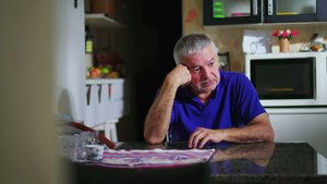 Bild mit traurig bklickenden Mann mit grauen Haaren, der alleine an einem Küchentisch sitzt und seinen Kopf auf eine Hand aufgestützt hält