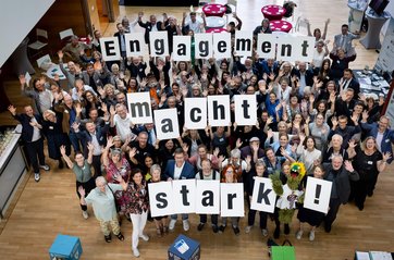 Bild von oben in einen Saal mit vielen lachenden und winkenden Menschen die Buchstabenschilder in die Höhe halten, sodass zu lesen ist: Engagement macht stark!