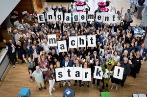 Bild von oben in einen Saal mit vielen lachenden und winkenden Menschen die Buchstabenschilder in die Höhe halten, sodass zu lesen ist: Engagement macht stark!