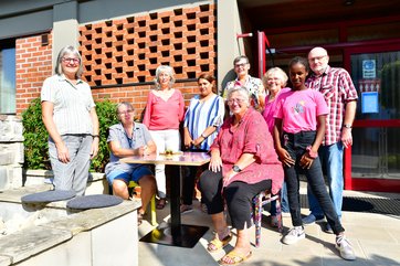 Bild mit sieben Frauen, einem Mann und einem Mädchen, die lachend vor einem Eingang stehen und sitzen.