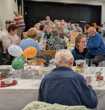 Viele gedeckte und geschmückte Tische, an denen Männer und Frauen unterschiedlichen Alters in einem größeren Raum sitzen, sich unterhalten, essen und trinken