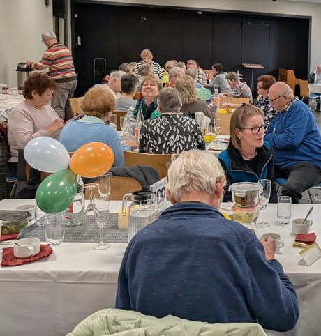 Viele gedeckte und geschmückte Tische, an denen Männer und Frauen unterschiedlichen Alters in einem größeren Raum sitzen, sich unterhalten, essen und trinken