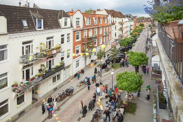 Mit buten Wimpeln und Luftballons geschmückter Straßenzug, Menschen stehen zusammen und feiern.