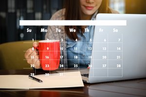 Monatskalender im Vordergrund, im Hintergrund sitzt Frau mit Tasse vor dem Computerbildschirm
