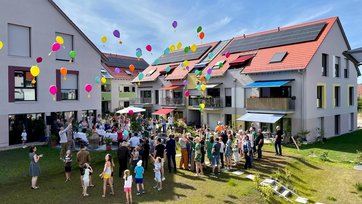 Menschen jeden alters stehen in Sommerkleidung auf einem Rasen vor einem Wohngebäudeensemble und lassen bei einer Feier bunte Luftballons steigen