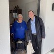 Mann steht mit Stoffbeutel an einer Haustür, die von einer lachenden älteren Frau am Rollator geöffnet wurde.