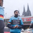 Bild mit sechs Männern, die hellblaue Radjacken mit der Aufschrift Muslime für Frieden tragen. Im Hintergrund ein Zug vor dem Kölner Dom