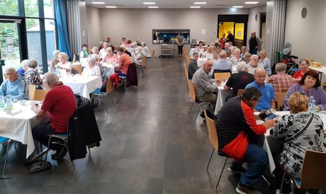 Bild mit Blick in einem Saal in dem viele Senioren und Seniorinnen an gedeckten Gruppentischen beim Essen sitzen.