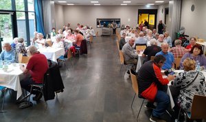 Bild mit Blick in einem Saal in dem viele Senioren und Seniorinnen an gedeckten Gruppentischen beim Essen sitzen.