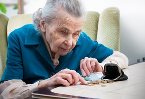 Alte Frau sitzt gebeugt im Sessel am Tisch vor geöffnetter Gelbörse und  zählt das wenig Münzgeld mit einer Hand in der andern Hand hält Sie einen Fünf-Euro-Schein