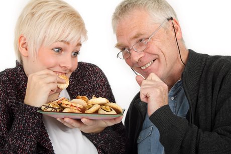 Ein Bild mit einer Frau und einem Mann. Die Frau hält einen Teller mit Plätzchen in der Hand und beißt in ein Plätzchen, ein älterer Mann mit Brille greift ein Plätzchen, beide lachen