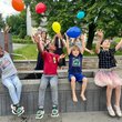Kinder siten lachend auf einer Bank im Freien und spielenm mit bunten Luftballons
