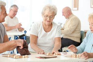 Ein alter Mann und zwei alte Frauen spielen ein Spiel am Tisch, im Hintergrund spielen zwei alte Männer ein Kartenspiel