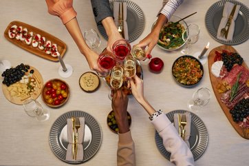fünf Hände unterschiedlicher Hautfarbe stoßen mit getränken über einem Tisch mit Mahlzeiten aus verschiedenen Ländern an