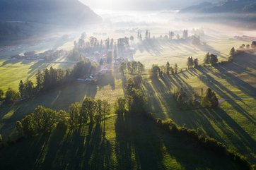 Dorf in Sonnestrahlen umgeben von Wiesen und Bachläufen in einem bewaldeten Tal.