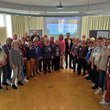 Gruppenbild mit vielen Männern und Frauen unterschiedlichen Alters, im Hintergrund eine Präsentation an der Wand.