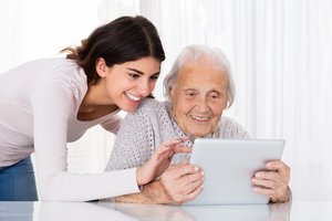 Junge Frau schaut alter Frau über die Schulter bei der Nutzung eines Tablets.