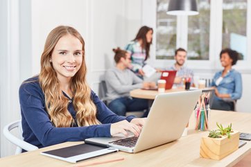 Junge Frau sitzt lächelnd an einem Laptop und arbeitet. Im Hintergrund sitzen vier junge Personen an einem runden Arbeitstisch zusammen.