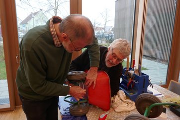 Bild mit zwei älteren Männern, die eine rote Kinderschubkarre auf einem Tisch reparieren