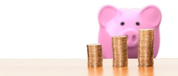 rosa Sparschweindose steht hinter 3 ansteigenden Geldmünzenstapeln auf einem Tisch