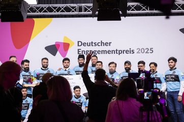 Gruppenfoto von Männern, die eine hellblaue Fahrradjacke tragen mit der Aufschirft Muslime für Frieden. Im Hintergrund das Logo Deutscher Engagementpreis 2025, im Vordergrund Publikum und Fotografen.