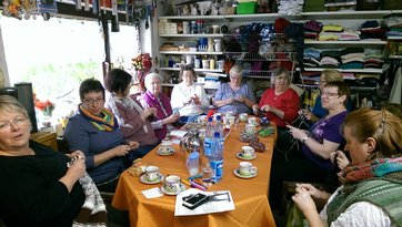 Elf Frauen sitzen häkelnd und strickend in einem Wollladen an einer großen Kaffeetafel