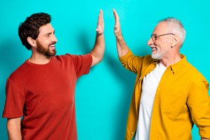 Bild mit einem jungen und alten Mann, die lachend und mit erhobenen Armen ihre Handflächen zusammenschlagen. 