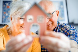 alte Frau und alter Mann halten Hausmodell in ihren Händen vor ihren Gesichtern und schauen mit einem Auge und lachend durch die Fenster