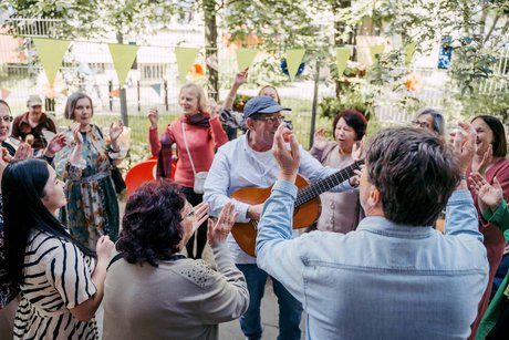 Ein Mann mit Gitarre steht im Freien inmitten einer Gruppe von Menschen die lachen tanzen und in die Hände klatschen
