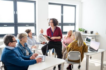 Lehrerin sitzt in einer Gruppe von Senior*innen, die an einer Klasse zur Computerfortbildung teilnehmen