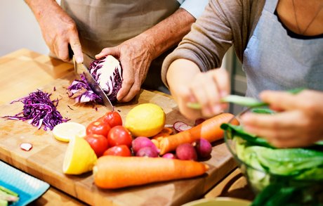Hände eines alten Mannes schneidet mit Messer Radicciosalat auf einem hölzernen Schneidebrett, auf dem auch Tomaten, Zitronen, Möhren und Radieschen liegen. Die Hände einer alten Frau legen grüne Salatblätter in eine Glasschüssel