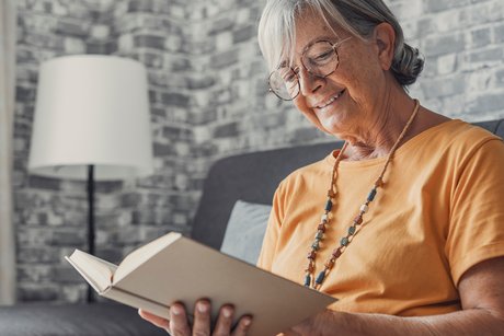 ältere Frau sitzt auf Sofa, hält Buch in der Hand und lacht