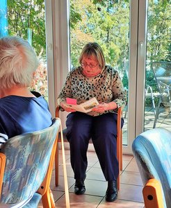 Frau über Buch gebeugt beim Vorlesen vor einer Gruppe älterer Menschen