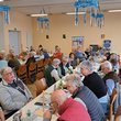 an langen Tischtafeln die bayerisch blau-weiß geschmückt sind sitzen Senioren beim Speisen und Getränken. In der Halle hängen einige blau-weiße Kränze von der Decke