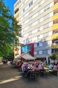 Menschen sitzen an Tischen und Banken vor einem Hochhaus bei blauem Himmel.