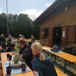 Bild mit Blockhütte auf einer Wiese, davor sitzen Menschen unterschiedlichen Alters auf Bierbänken an Tischen mit Kaffeetassen und Kuchen