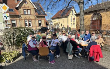 Bild mit einer Gruppe von Menschen die auf Stühlen an zwei gedeckten Tischen bei blauem Himmel im Freien in einem Ort sitzen.