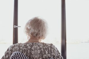 Alte Frau mit weißen Haaren sitzt auf einem Stuhl und blickt mit dem Rücken zum Betrachter durch ein Fenster nach draußen