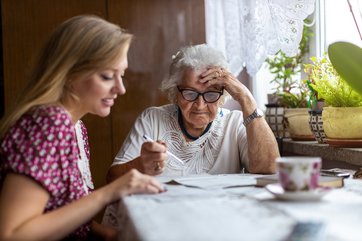 Junge Frau mit langen Haaren sitzt am Tisch mit alter Frau mit grauen Haaren und Brille. Auf dem Tisch Papierunterlagen und eine Porzellantasse. Junge Frau hilft Älterer beim Ausfüllen der Formulare