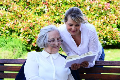 Ältere Frau sitzt auf einer Bank im Freien und schaut in ein Buch, das eine jüngere Frau in der Hand hät, die sich über die Rückenlehne  der Bank beugt. Beide lächeln