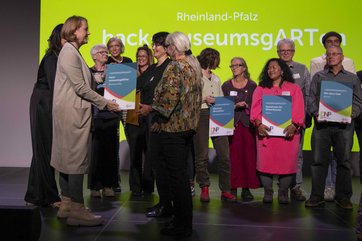 Bild mit der Ministerin die Urkunde an eine Frau übergibt. Auf der Bühne viele weitere Menschen mit Urkunden