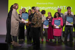Bild mit der Ministerin die Urkunde an eine Frau übergibt. Auf der Bühne viele weitere Menschen mit Urkunden