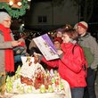 Abendliche Stimung, warm gekleidete Menschen auf dem Weihnachtsmarkt im Vordergrund das Lebkuchenhaus, ältere Frau überreicht jüngeren Frau ein Buch