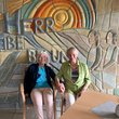 Zwei ältere Frauen sitzen lachend und Handhaltend in Stühlen vor einem großen Wandmosaik.