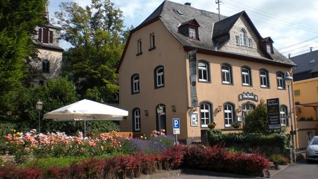 Bild mit altem schiefergedeckten Gebäude, davor ein Rosengarten mit Sonnenschirm