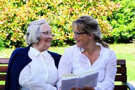 Foto einer jüngeren Frau, die mit einer älteren Frau im Park auf einer Bank sitzt und ein Buch in der Hand hält und der alten Frau präsentiiert, die Frauen lachen sich an.