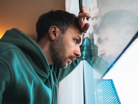 Bild mti einem mittelalten Mann, der traurig schaut und der mit Hand und Kopf an einem Fenster lehnt